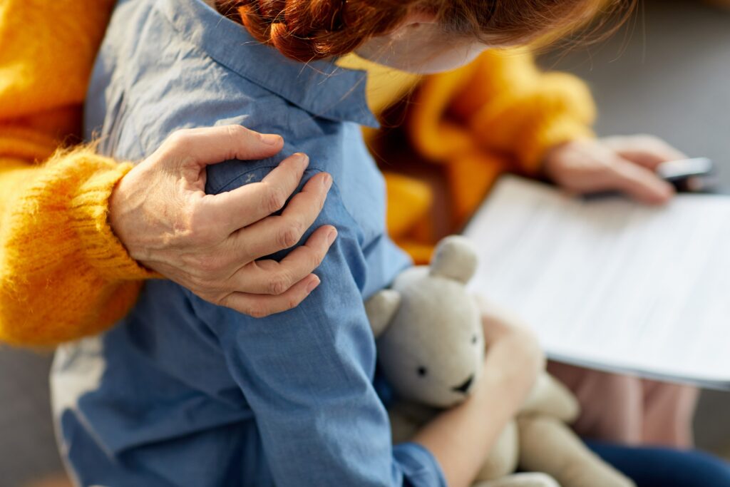 A child holds a teddy bear with an adults hand on their shoulder to comfort them