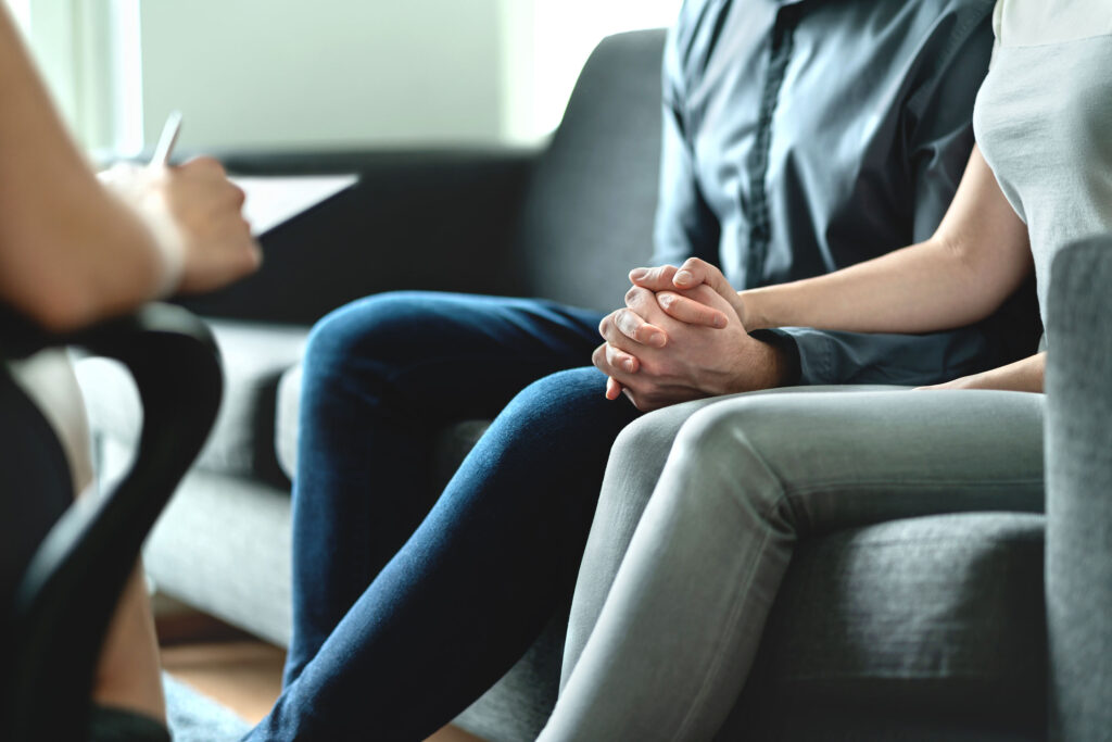 A couple sit on a grey chair during a couple therapy session.